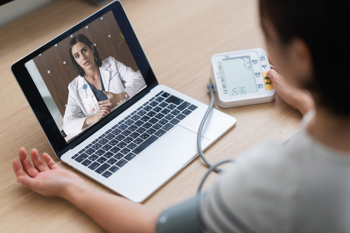 A woman is measuring her blood pressure while consulting with a doctor via telemedicine video call on a laptop at home