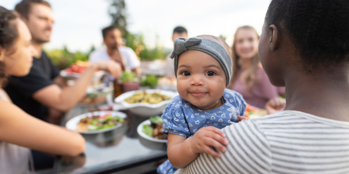A multiethnic group of young friends enjoy good food and conversation together on a terrace outside on a summer evening. The focus is on an adorable young girl who's smiling at the camera while mom holds her.