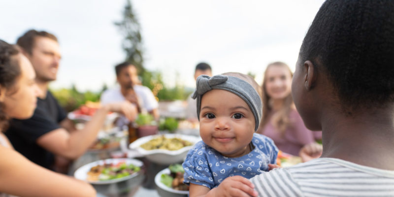 A Multiethnic Group Of Young Friends Enjoy Good Food And Conversation Together On A Terrace Outside On A Summer Evening. The Focus Is On An Adorable Young Girl Who's Smiling At The Camera While Mom Holds Her.