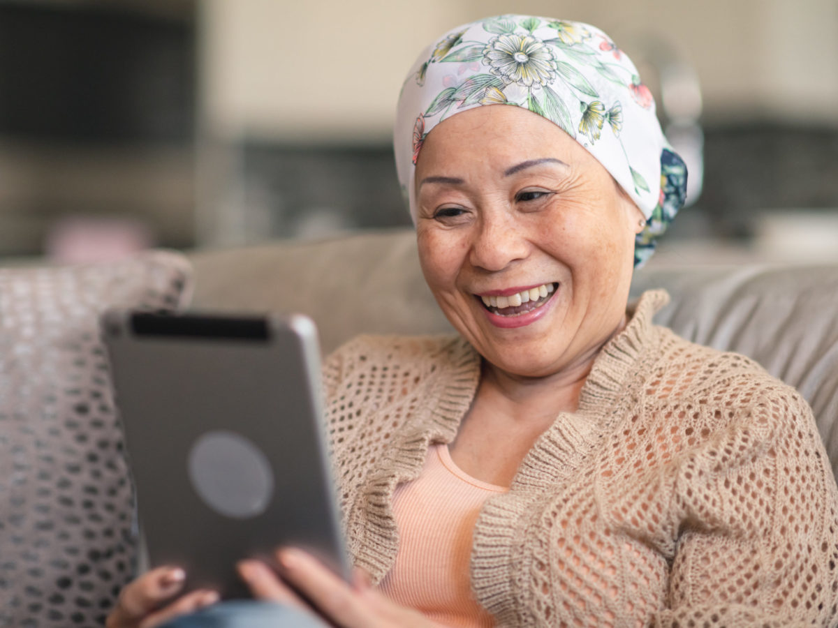 A Senior Adult Woman Of Chinese Descent Has Cancer. The Happy Woman Is Using A Digital Tablet To Video Message Friends And Family. She Is Spending Time At Home. The Woman Is Smiling At The Screen. She Is Wearing A Bandana To Hide Her Hair Loss From Chemotherapy Treatment.