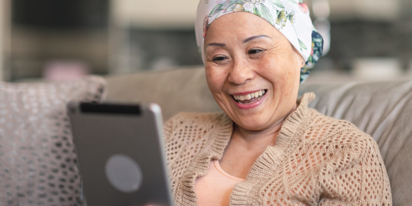 A Senior Adult Woman Of Chinese Descent Has Cancer. The Happy Woman Is Using A Digital Tablet To Video Message Friends And Family. She Is Spending Time At Home. The Woman Is Smiling At The Screen. She Is Wearing A Bandana To Hide Her Hair Loss From Chemotherapy Treatment.