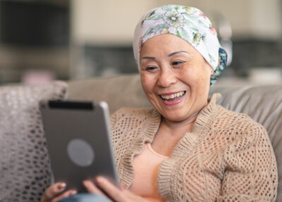 A senior adult woman of Chinese descent has cancer. The happy woman is using a digital tablet to video message friends and family. She is spending time at home. The woman is smiling at the screen. She is wearing a bandana to hide her hair loss from chemotherapy treatment.
