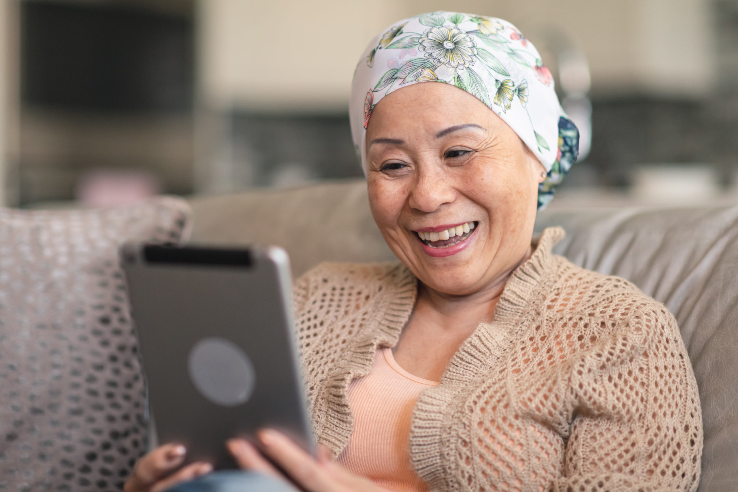 A senior adult woman of Chinese descent has cancer. The happy woman is using a digital tablet to video message friends and family. She is spending time at home. The woman is smiling at the screen. She is wearing a bandana to hide her hair loss from chemotherapy treatment.