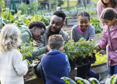 A mature African-American man teaching a group of five multiracial children how to grow vegetable plants in a community garden. He is holding a tray of potted plants, kneeling down so the girls and boys, 4 to 10 years old, can see them as he talks. The boy standing beside him is his son.