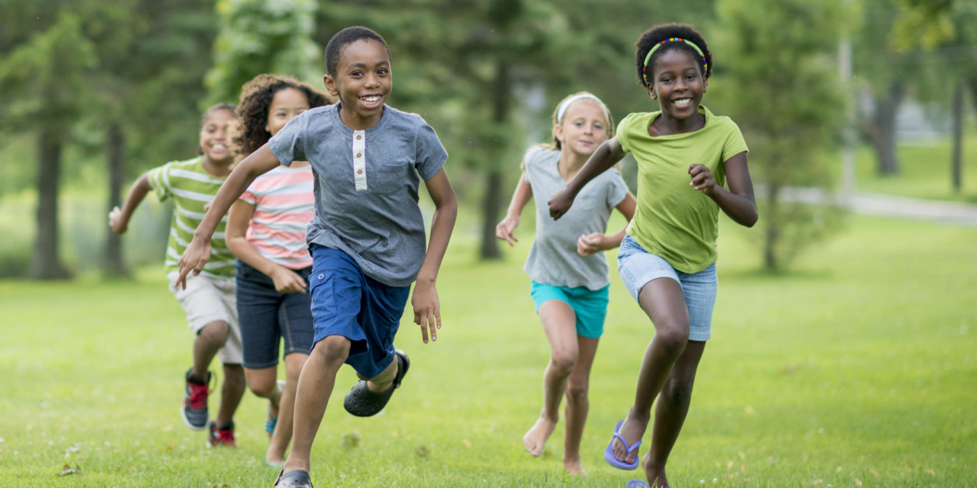 A Multi-ethnic Group Of Elementary Age Students Are Playing Tag At The Park During Recess. They Are Happily Chasing Each Other Through The Grass On A Sunny Day.