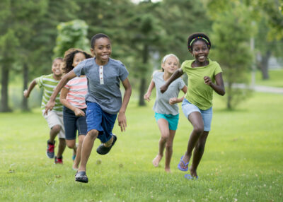 A multi-ethnic group of elementary age students are playing tag at the park during recess. They are happily chasing each other through the grass on a sunny day.