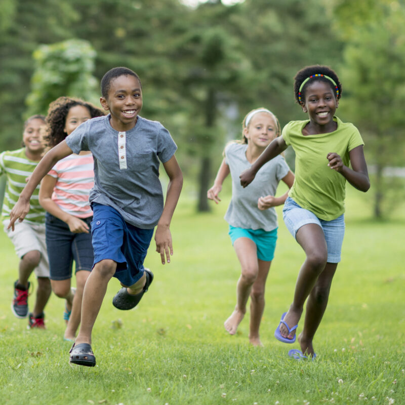A multi-ethnic group of elementary age students are playing tag at the park during recess. They are happily chasing each other through the grass on a sunny day.