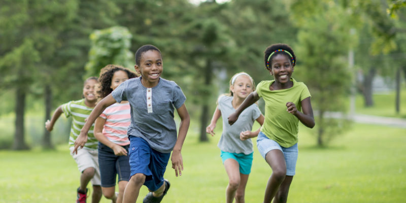 A Multi-ethnic Group Of Elementary Age Students Are Playing Tag At The Park During Recess. They Are Happily Chasing Each Other Through The Grass On A Sunny Day.