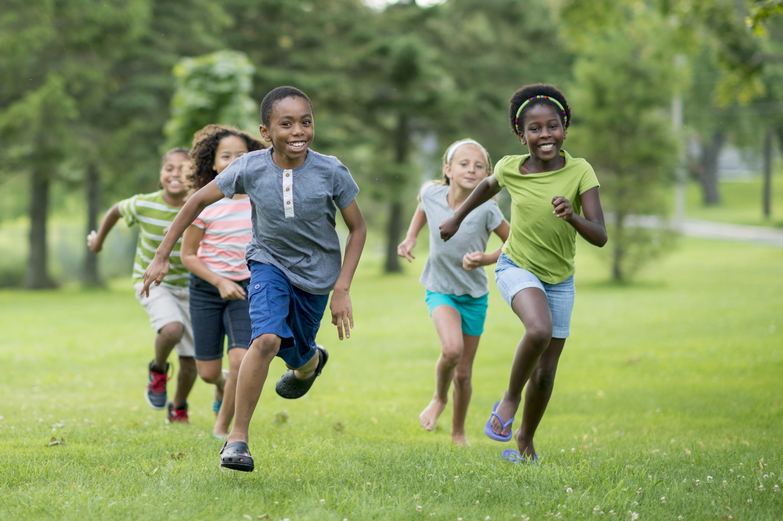 A multi-ethnic group of elementary age students are playing tag at the park during recess. They are happily chasing each other through the grass on a sunny day.