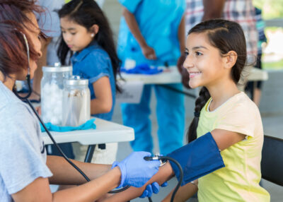 Cheerful preteen Hispanic girl having her blood pressure checked by a female African American mid adult volunteer nurse at an outdoor health fair. Smiling Hispanic young girl at a medical expo getting her blood pressure checked by African American volunteer nurse.