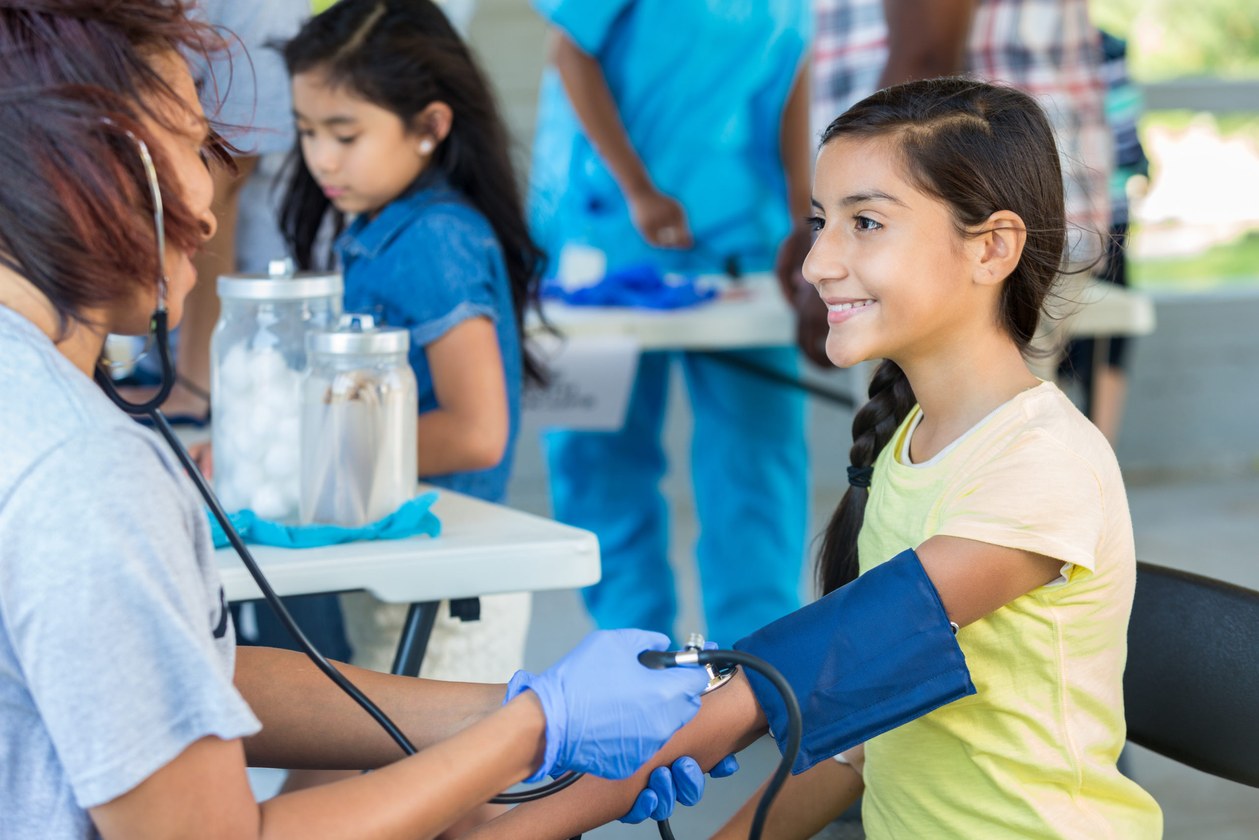Cheerful preteen Hispanic girl having her blood pressure checked by a female African American mid adult volunteer nurse at an outdoor health fair. Smiling Hispanic young girl at a medical expo getting her blood pressure checked by African American volunteer nurse.