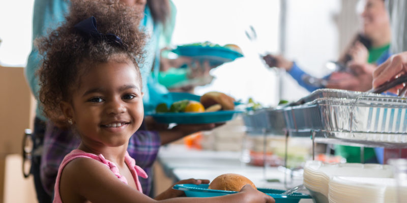 Cheerful Black little girls smiles while in line in a soup kitchen. She is holding a plate full of healthy food. Her family is in line behind her.