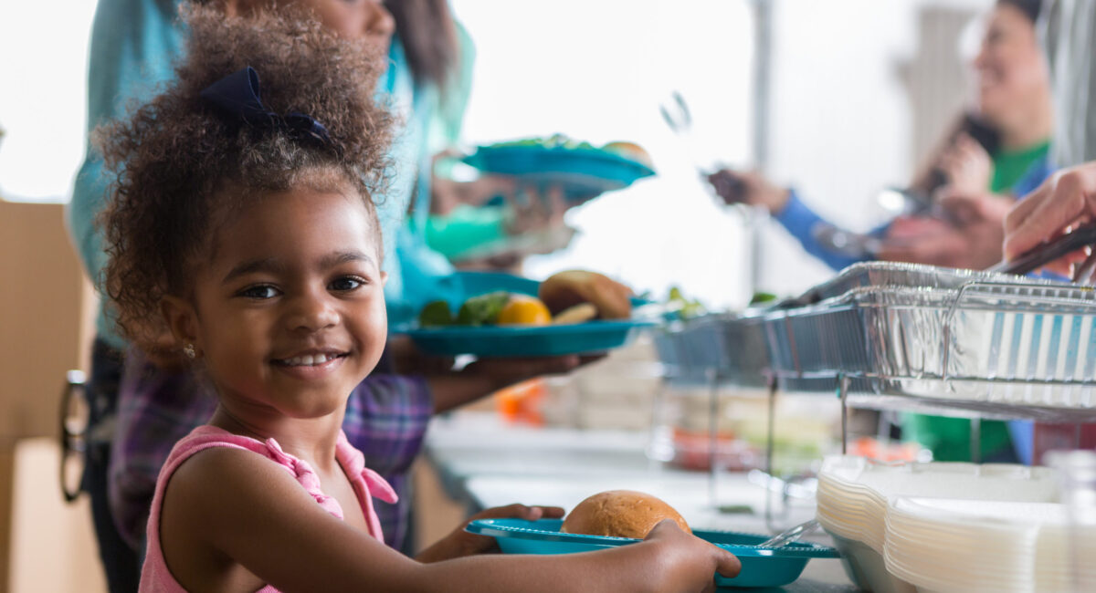 Cheerful Black little girls smiles while in line in a soup kitchen. She is holding a plate full of healthy food. Her family is in line behind her.