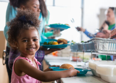 Cheerful Black little girls smiles while in line in a soup kitchen. She is holding a plate full of healthy food. Her family is in line behind her.