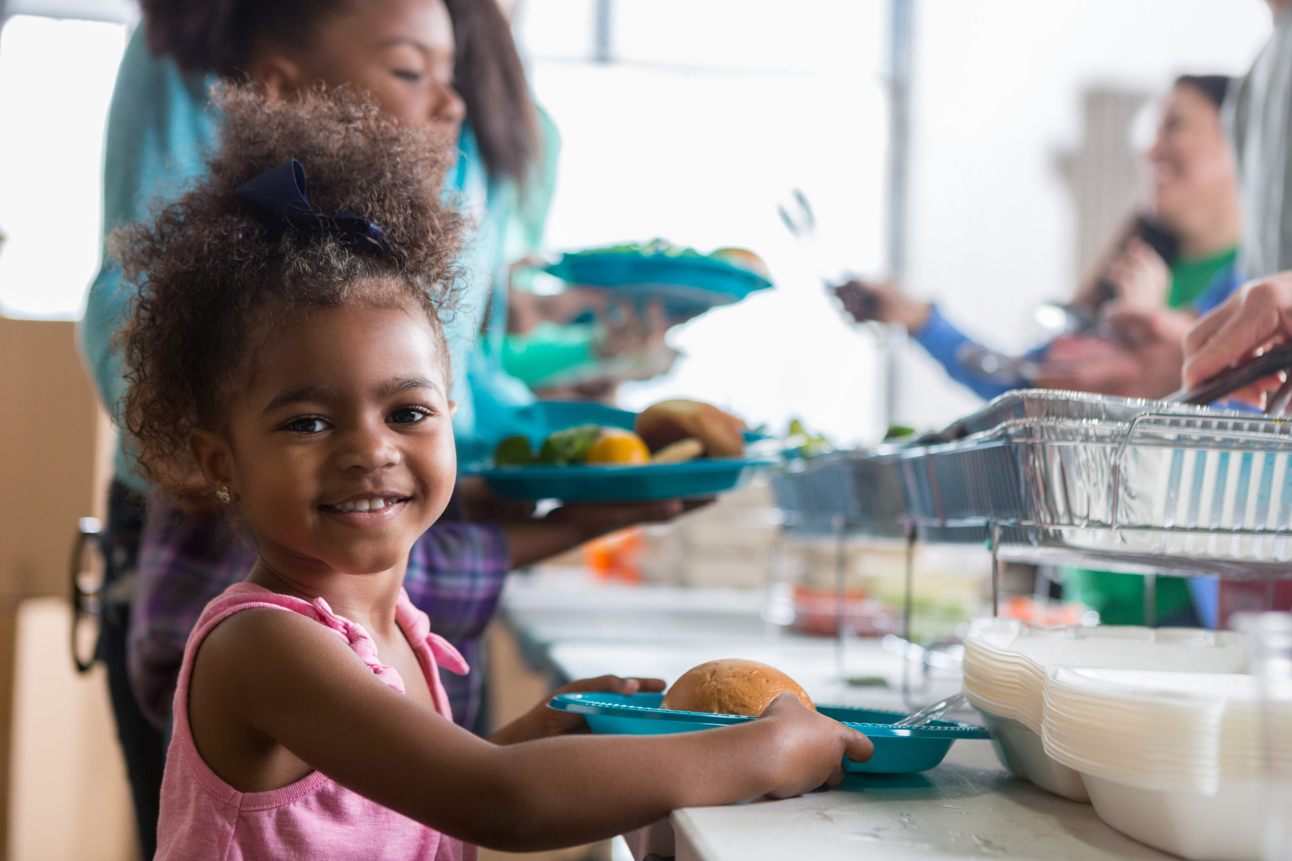 Cheerful Black little girls smiles while in line in a soup kitchen. She is holding a plate full of healthy food. Her family is in line behind her.