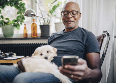 Smiling retired senior male using smart phone while sitting with dog in room at home