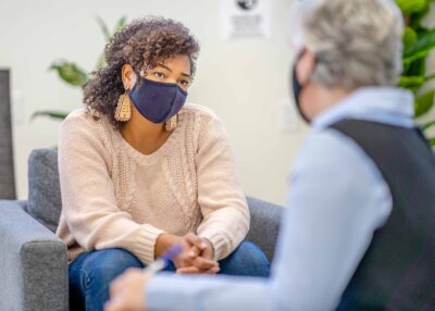 A young female of African ethnicity is seated across her therapist at a therapy session. She appears to be listening to her therapist.
